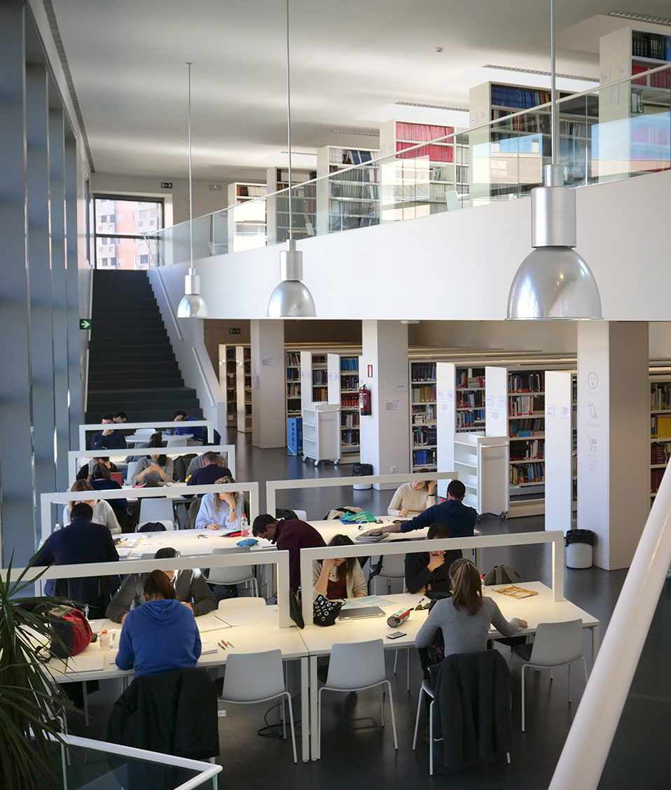 Interior de la biblioteca biosanitaria donde se pueden ver estanterías repletas de libros, estudiantes en las mesas de estudio y dos escaleras que llevan a la planta superior